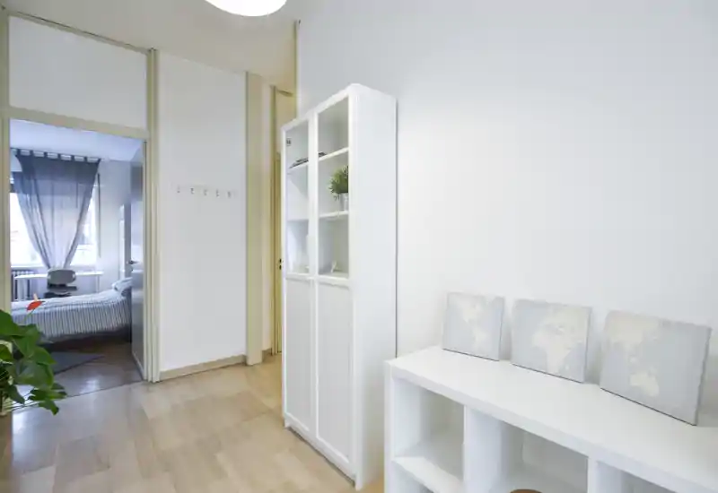 Vista luminosa y minimalista del dormitorio visible a través de una puerta abierta con cama bien hecha, cortinas ligeras y un pequeño escritorio junto a la ventana; en primer plano un armario blanco y estanterías.