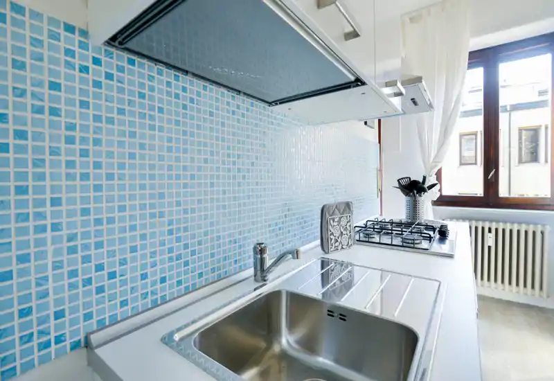 Bright, modern kitchen corner with a stainless steel sink, gas hob and light blue mosaic backsplash, well lit by a nearby window.