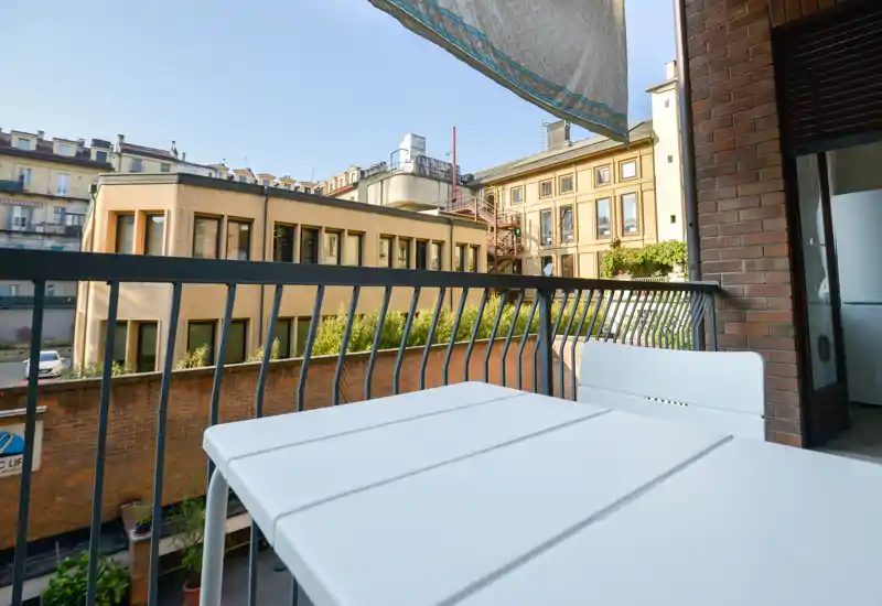 Balcone cittadino soleggiato con piccolo tavolo e sedia bianchi, ringhiera in metallo e vista sugli edifici vicini — luminoso e ideale per il caffè mattutino.