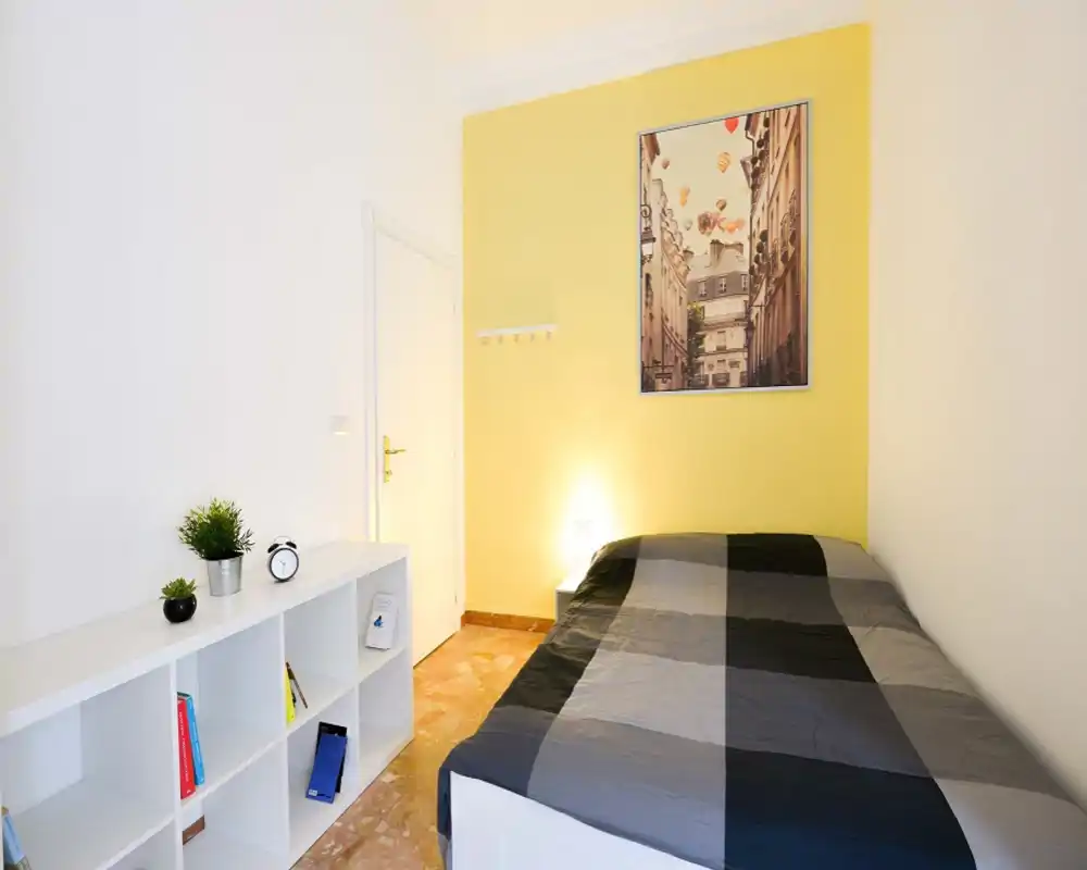 Compact, well-lit bedroom with a double bed dressed in a gray patterned duvet, a bedside lamp and framed wall art on a warm yellow accent wall. A low white shelving unit and small decorative plants complete the tidy layout.