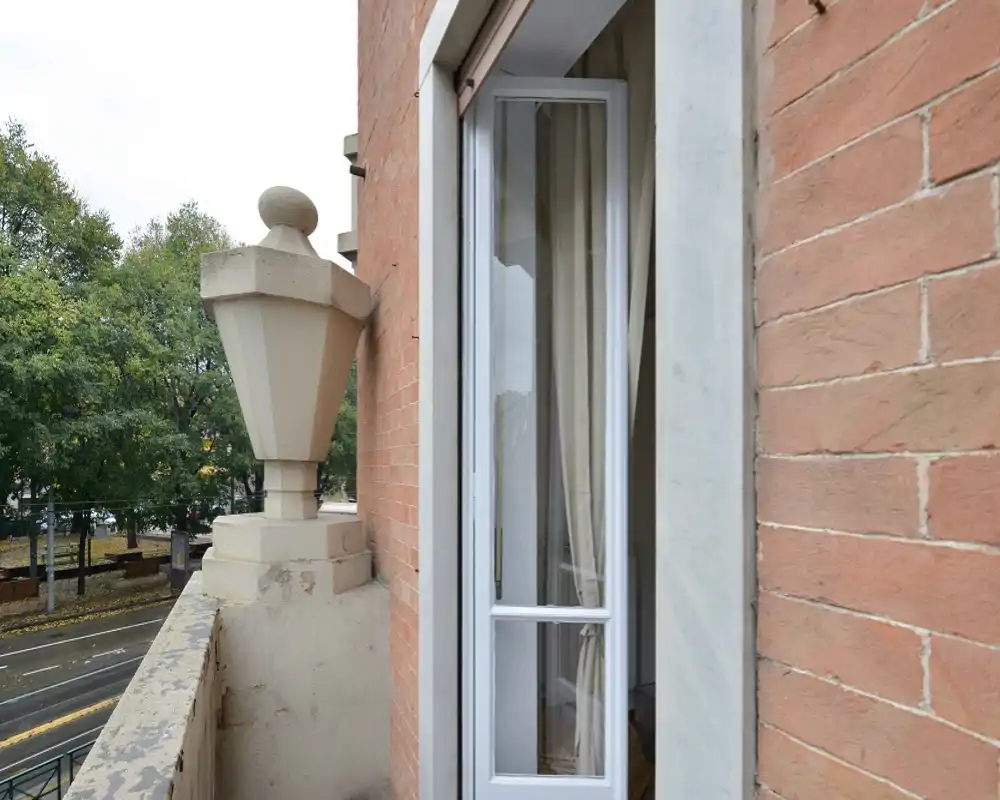 Small exterior balcony with a stone balustrade and a view over a tree-lined street; a partially open window with curtains connects the balcony to the interior.
