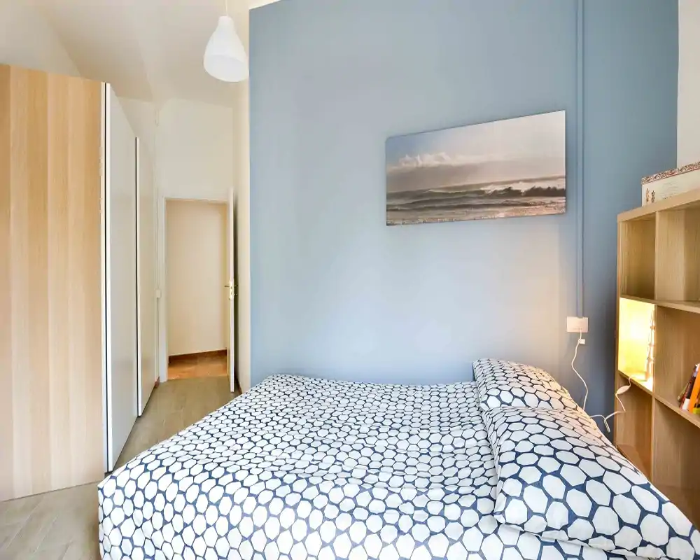 Camera da letto matrimoniale luminosa e ben arredata con letto a pattern, parete accento azzurra e armadi integrati. Spazio pulito, accogliente e ben illuminato, ideale per annunci di affitto.
