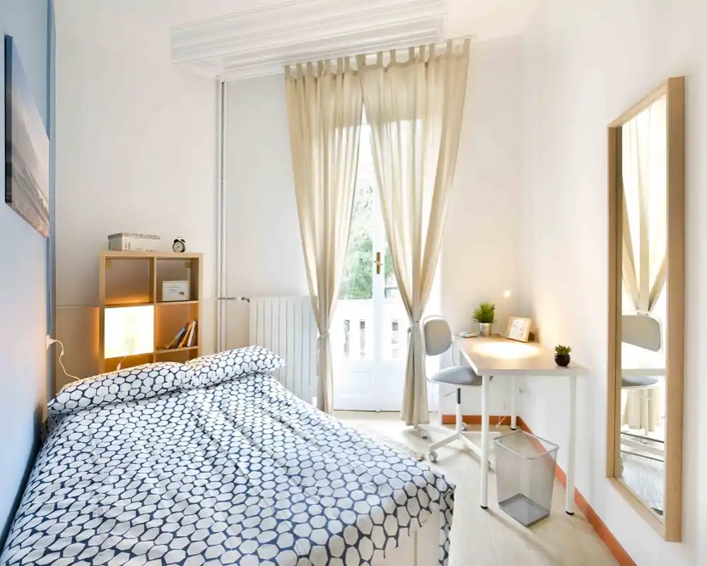 Camera da letto luminosa e ben arredata con letto matrimoniale, biancheria morbida, una piccola scrivania vicino alla finestra e uno specchio a figura intera. La luce naturale entra attraverso tende leggere, rendendo l'ambiente arioso e accogliente.
