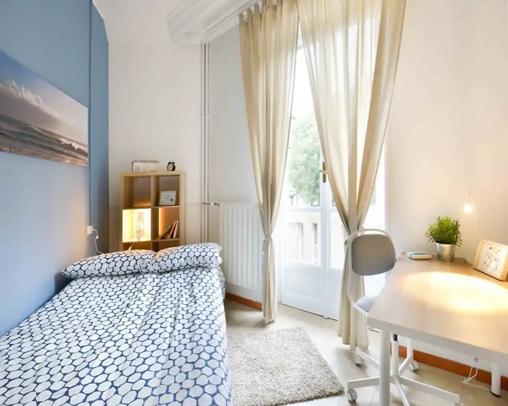 Camera da letto luminosa e ordinata con letto a fantasia, scrivania vicino alla porta finestra e luce naturale morbida — ideale per studenti o professionisti.