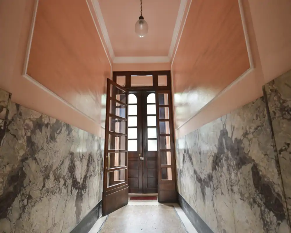 Symmetrical entrance hallway with decorative marble lower walls, warm painted upper walls and a wooden double door with glass panels. Bright, welcoming passage ideal as the building's main entry.