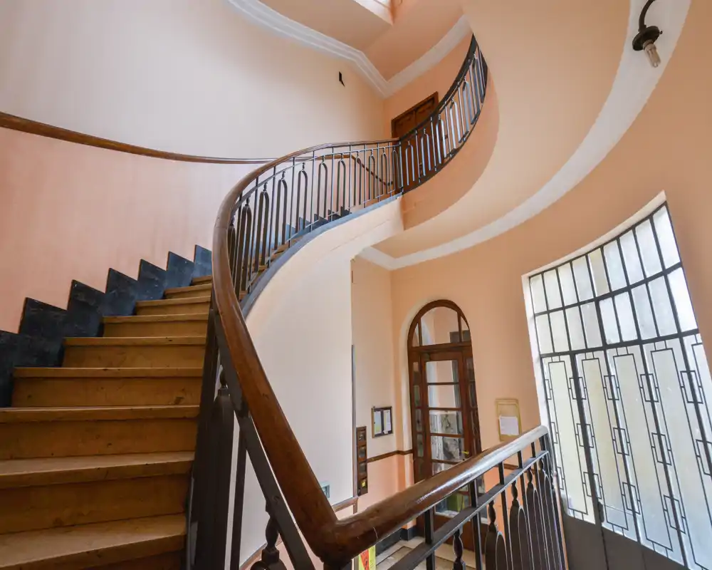 Bright, elegant entrance with a curved wooden staircase and tall window allowing generous natural light — ideal for a welcoming building lobby.