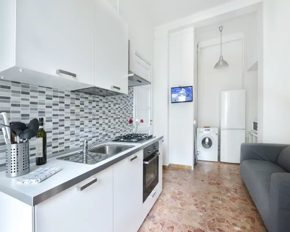 Compact, modern kitchenette with white cabinetry, gas cooktop, stainless sink, mosaic tile backsplash and a small living area visible; bright and neatly arranged for short-term rentals.