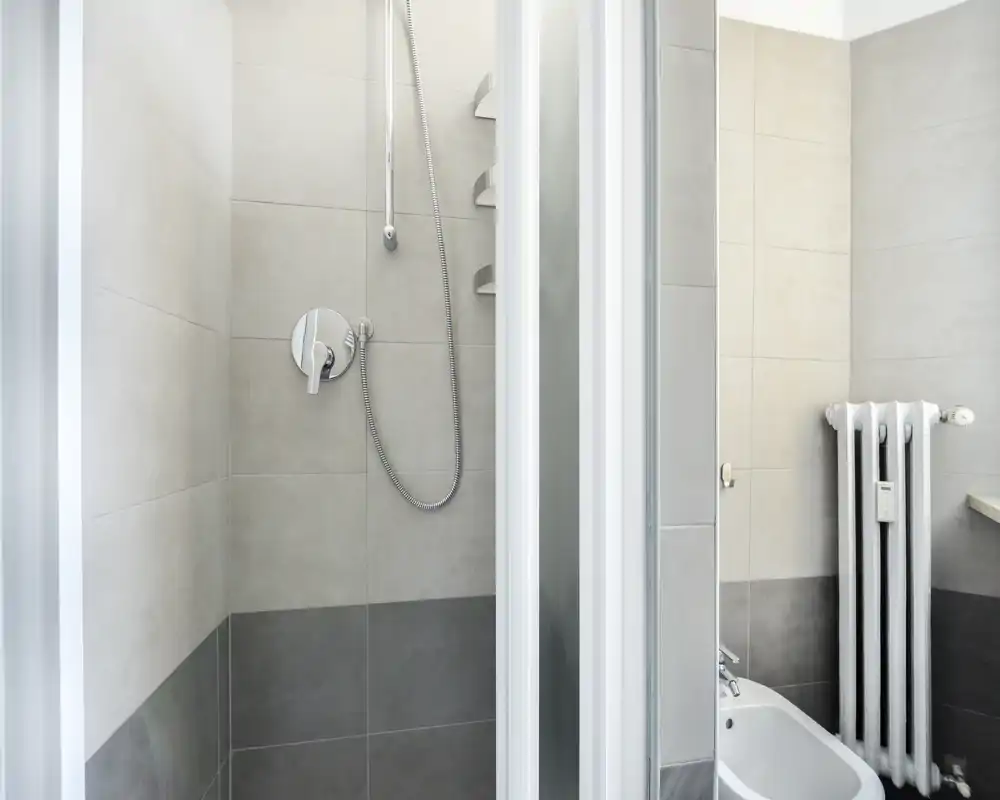 Modern tiled bathroom with a walk-in shower, visible shower controls and a bidet next to a vertical radiator; neutral grey-beige tones and clean, minimal styling.