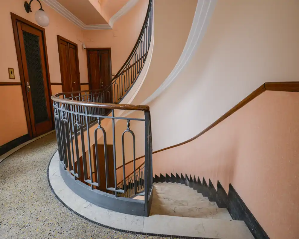 Bright, curved stairwell with decorative iron railing and terrazzo flooring — a welcoming communal entrance area with warm pastel walls and wooden doors.