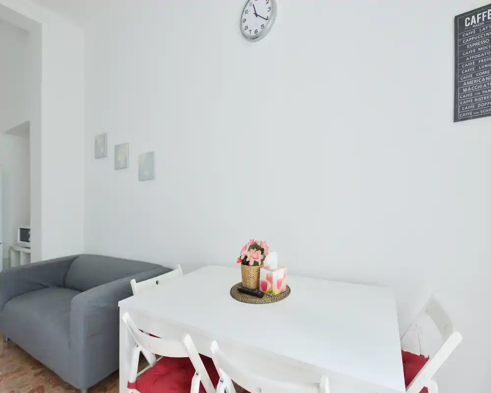 Bright, minimal living area with a white dining table and a compact grey sofa; simple decorative accents like a small centerpiece and wall clock give a welcoming feel.