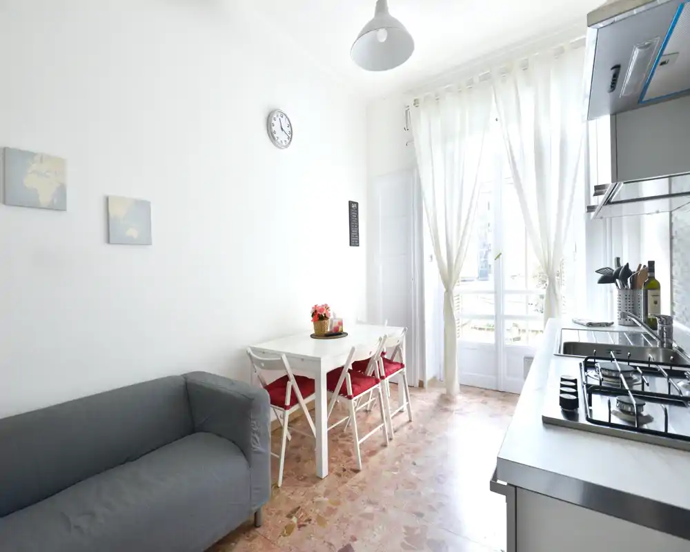 Bright, compact kitchen area with a small dining table and four chairs, modern gas hob and sink along the counter, and a cozy sitting corner next to large French doors letting in abundant daylight.