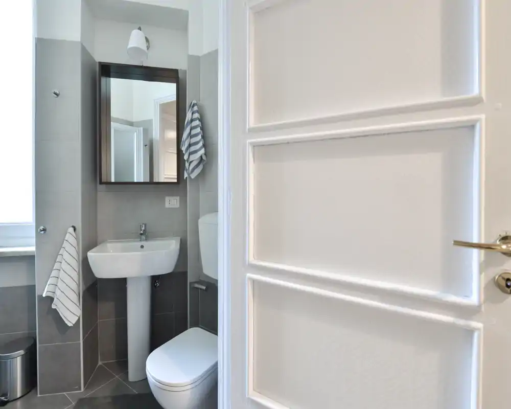 Small modern bathroom with a pedestal sink, wall-mounted mirror cabinet, toilet and striped hand towels; bright natural light from a window and neutral grey tiles.