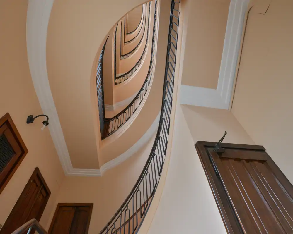 View looking up a curved interior stairwell and landing with decorative railings and wooden doors — a well-lit entrance / stair core with architectural detail.