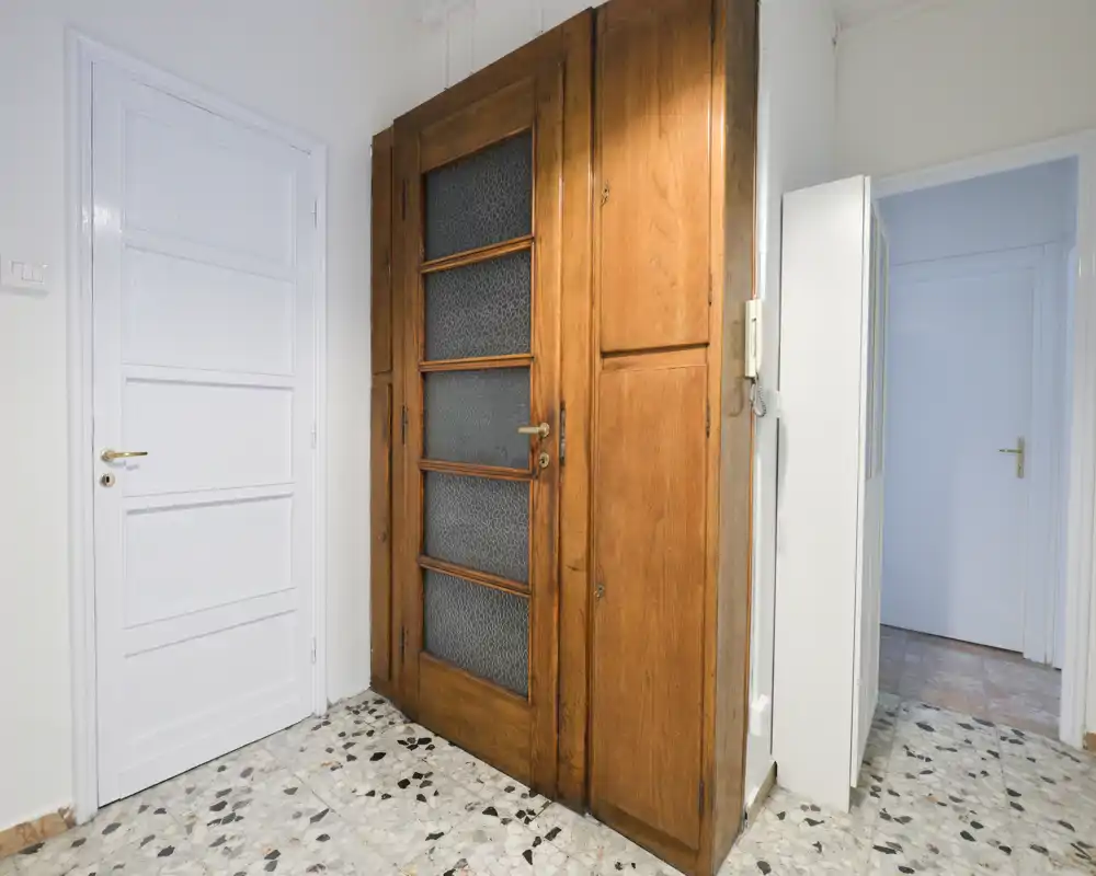 Bright entrance/foyer with a vintage wooden door and terrazzo floor; clean white walls and an adjacent hallway visible.