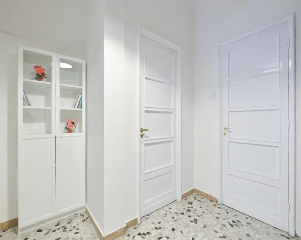 Bright, minimalist hallway entrance with two white interior doors and a slim glass-front cabinet; terrazzo floor and neutral walls create a clean welcoming entry.