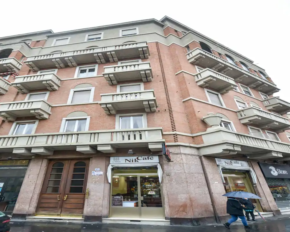 Exterior street-level view of a multi-story brick building with small shopfronts (NikCafé) at ground level and pedestrian activity on a rainy day.
