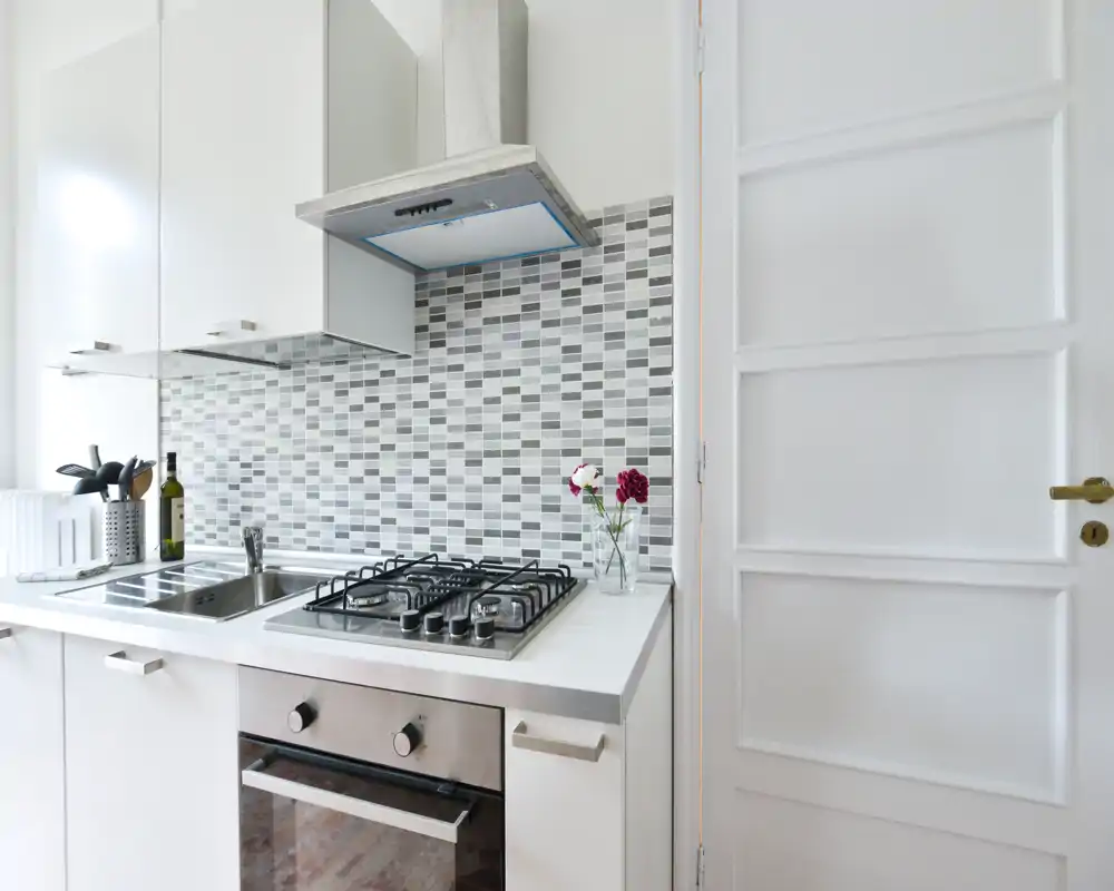 Bright modern kitchenette with a stainless steel gas hob, oven and sink against a stylish mosaic tile backsplash; neat white cabinetry and small decorative flowers create a fresh, welcoming feel.
