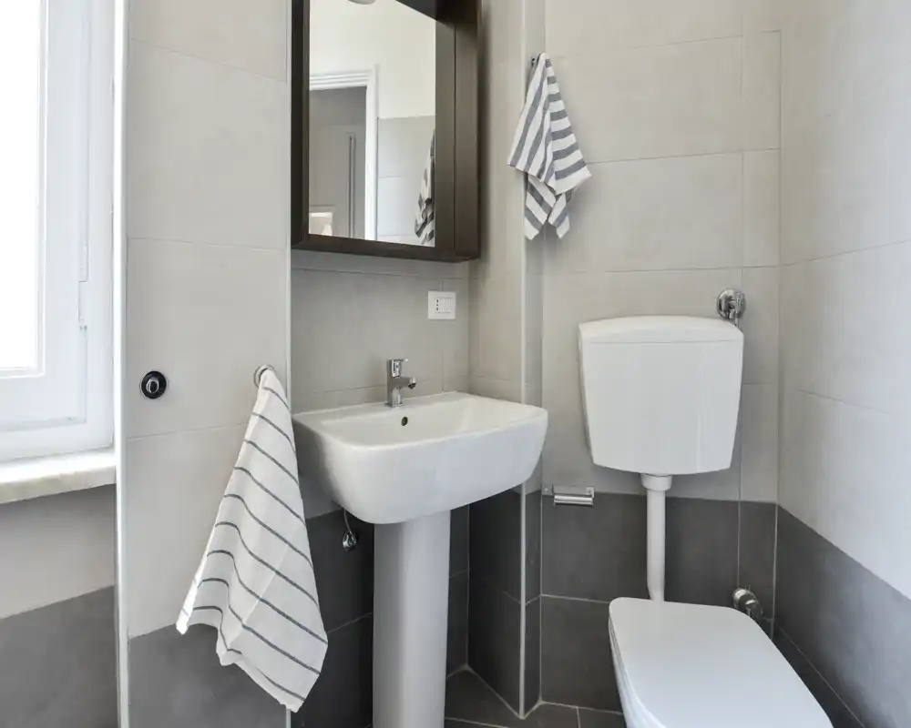 Compact, clean bathroom with a pedestal sink, toilet with high-mounted cistern and a mirrored medicine cabinet; neutral grey tiles and striped towels create a fresh, minimal look.