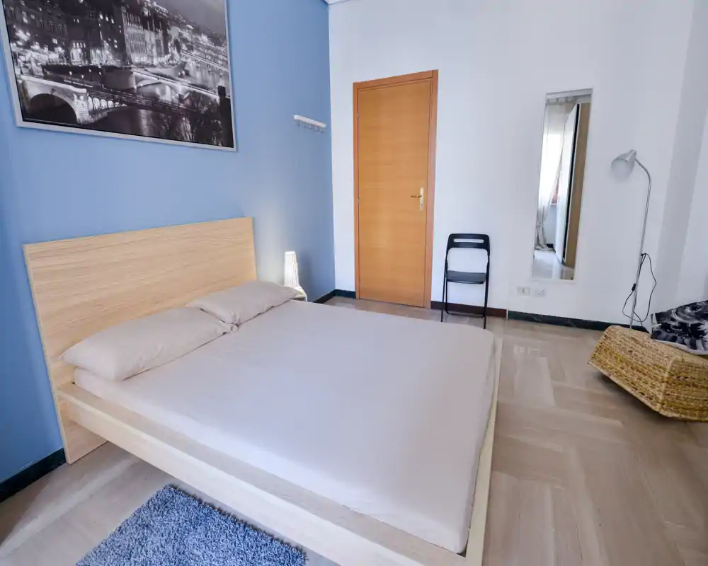 Bright, modern bedroom with a wooden platform bed, neutral bedding and soft lighting. The room features a blue accent wall, simple decor and a full-length mirror near the door.