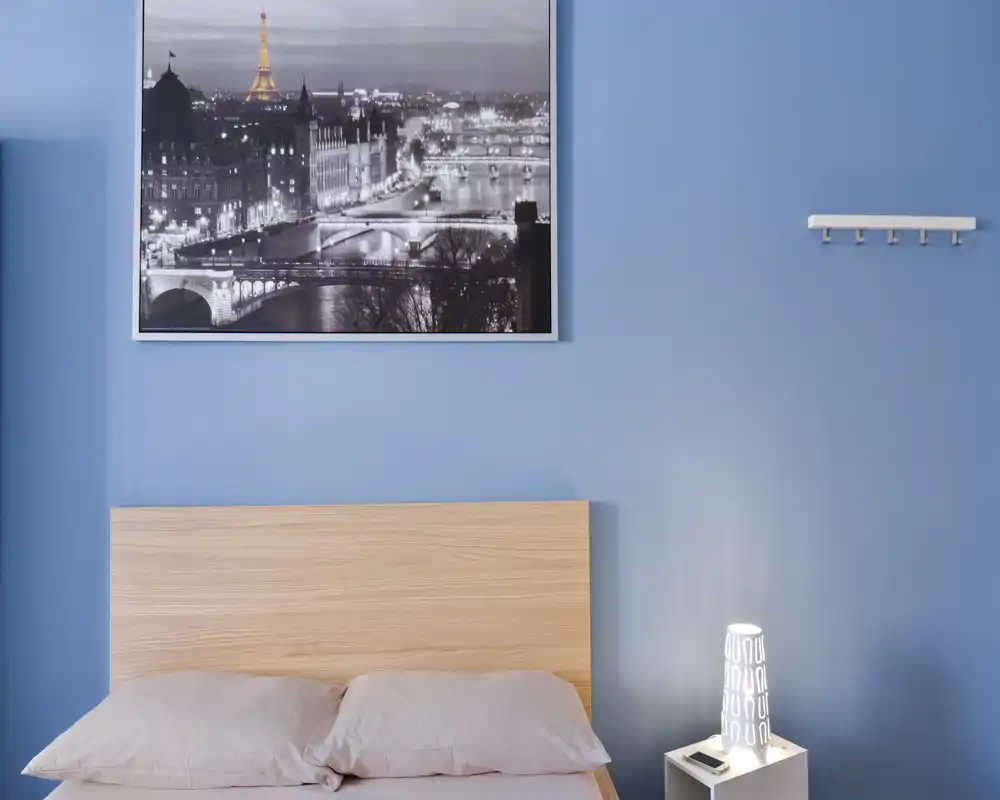 Bright, tidy bedroom with a light wood headboard, soft pillows and calm blue walls accented by a framed city photo and a bedside lamp.