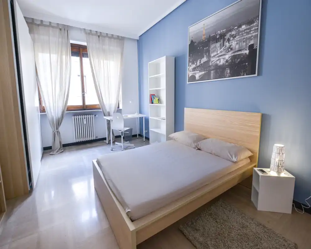 Bright, modern bedroom with a wooden platform bed, large window with sheer curtains, bedside lamp and a small desk — airy and well-lit space ideal for relaxing or working.