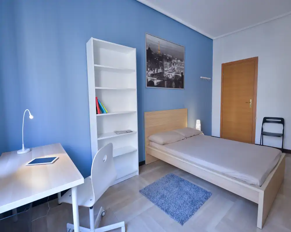 Bright, tidy bedroom with a double bed, simple wooden frame, bedside lamp and a study desk by a blue accent wall — ideal for a single occupant or couple seeking a minimalist space.