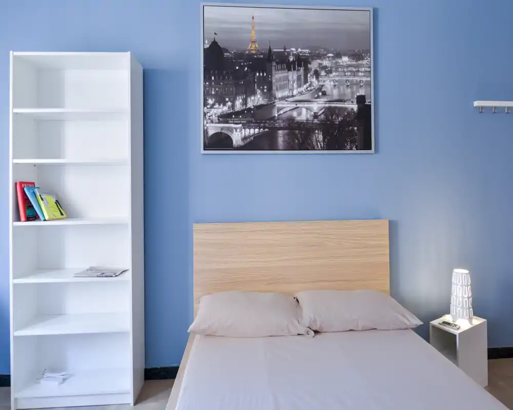 Bright, minimal bedroom with a wooden headboard, neutral bedding and a bedside lamp; a tall white bookshelf and framed cityscape make the space feel modern and airy.