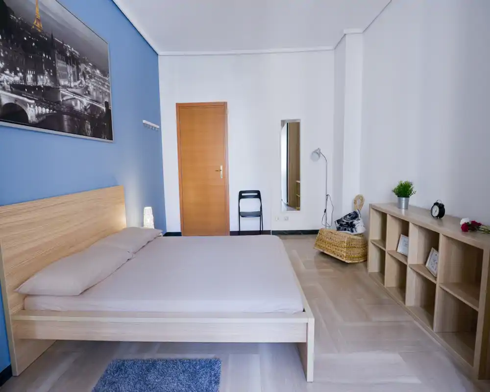 Spacious, well-lit bedroom with a modern wooden bed, soft linens, a bedside lamp and tasteful shelving — bright and welcoming for guests.