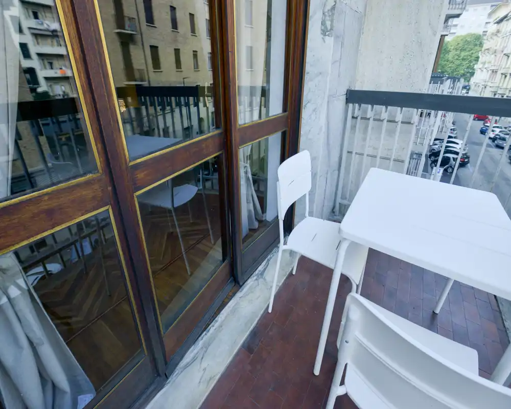 Pequeño balcón urbano con mesa y sillas blancas, suelo de baldosas y vista a la calle; ideal para el café de la mañana.