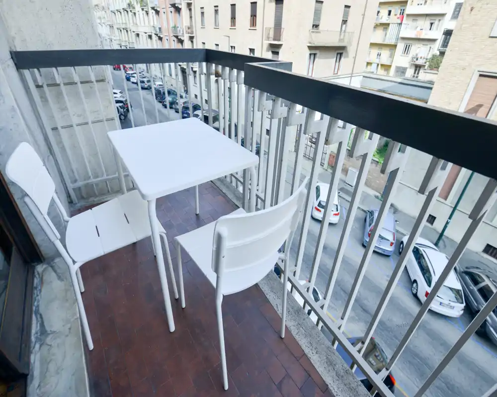 Balcón estrecho y soleado con una pequeña mesa blanca y dos sillas con vistas a una calle tranquila — ideal para el café de la mañana.