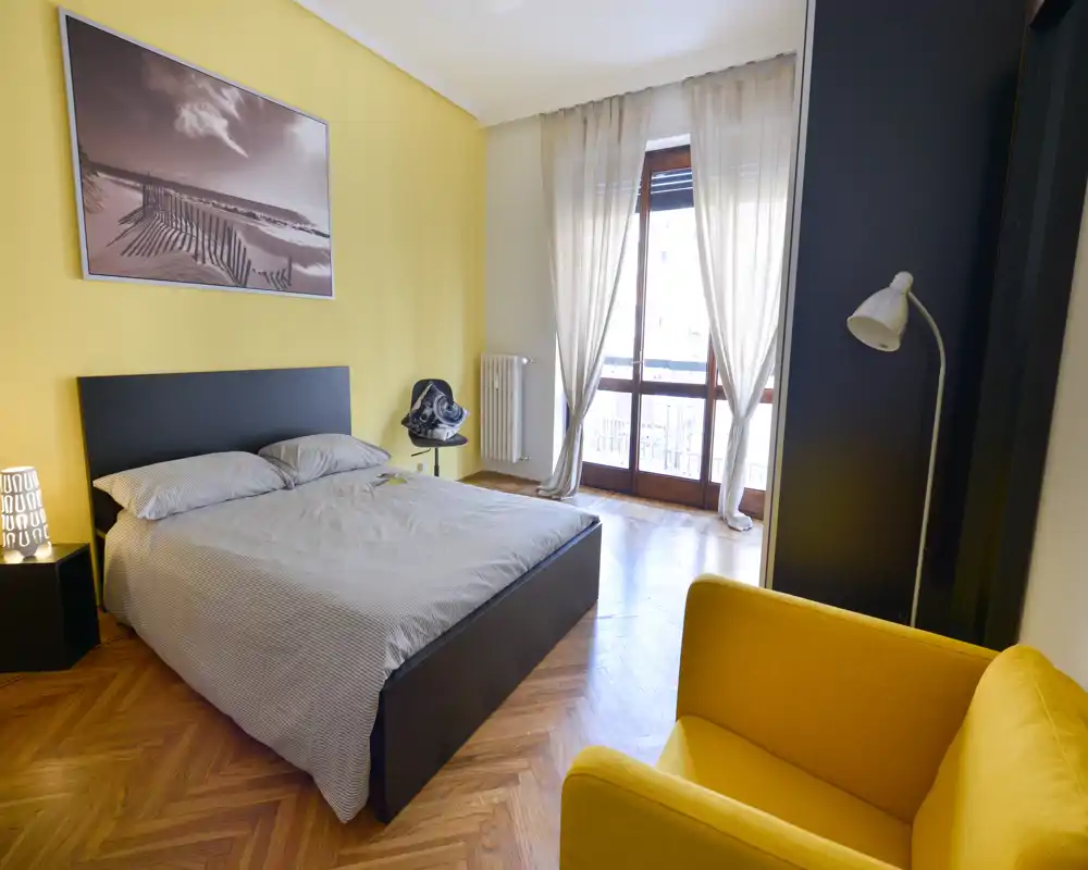 Dormitorio moderno y luminoso con cama doble frente a una pared amarilla, suelo de parquet y una gran ventana que aporta luz natural. Un sillón mostaza y mobiliario minimalista ofrecen una sensación acogedora y contemporánea.