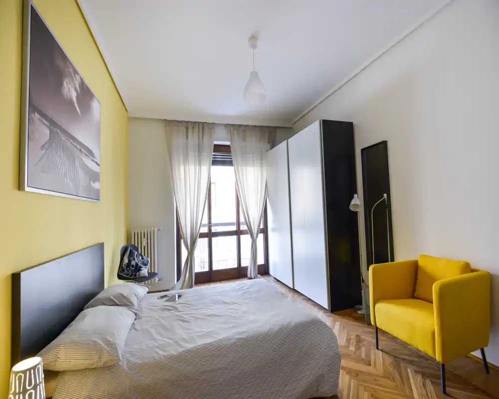 Dormitorio luminoso y bien decorado con una cama doble frente al balcón, armario moderno y un sillón amarillo. La luz natural y el suelo de madera crean un ambiente acogedor, perfecto para anuncios de alquiler.