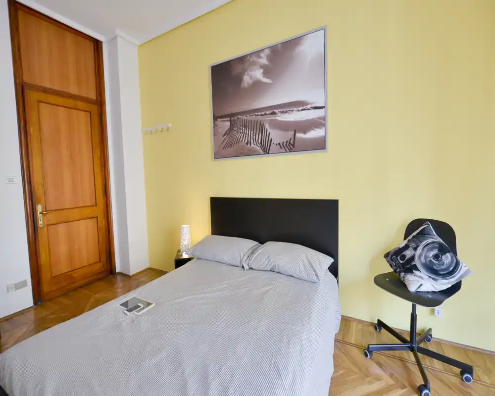 Dormitorio luminoso y de estilo sencillo con cama doble, lámpara de mesita, cuadro en la pared y una silla giratoria. La composición destaca la cama y el suelo de madera para una sensación acogedora.