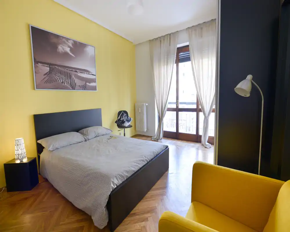 Dormitorio moderno y luminoso con cama doble, gran ventanal con cortinas traslúcidas, suelo de parquet en espiga y sillón amarillo; líneas limpias y luz natural que lo hacen agradable.