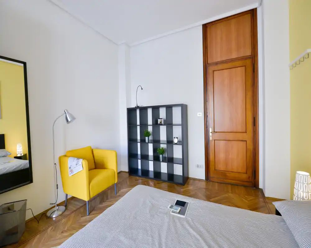 Dormitorio luminoso y con estilo, con la cama visible en primer plano, un acogedor sillón amarillo, estantería moderna y puerta de madera; suelo de parquet y luz natural crean ambiente cálido.