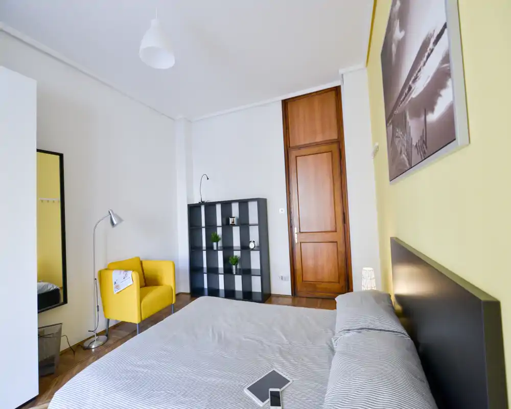 Dormitorio moderno y luminoso con cama grande, ropa de cama a rayas, un sillón amarillo y estantería — bien ambientado y espacioso.