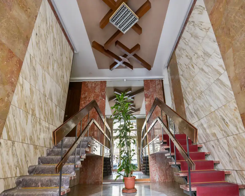 Vestíbulo de entrada señorial con escalera doble simétrica, paredes revestidas de mármol, vigas decorativas en el techo y planta central en maceta; suelos pulidos y barandillas ofrecen una primera impresión elegante.