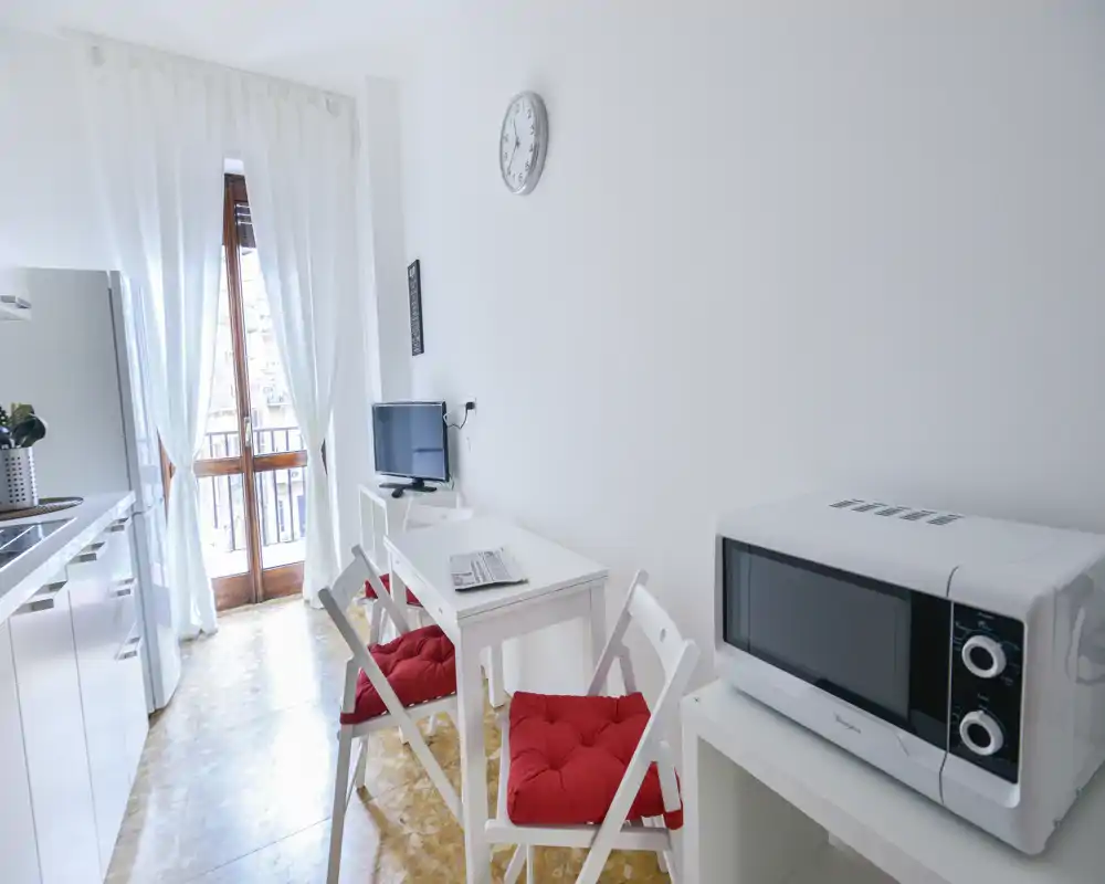 Cocina moderna y compacta con zona de comedor, mobiliario blanco, microondas y luz natural desde la puerta al balcón — ideal para estancias cortas y acogedoras.