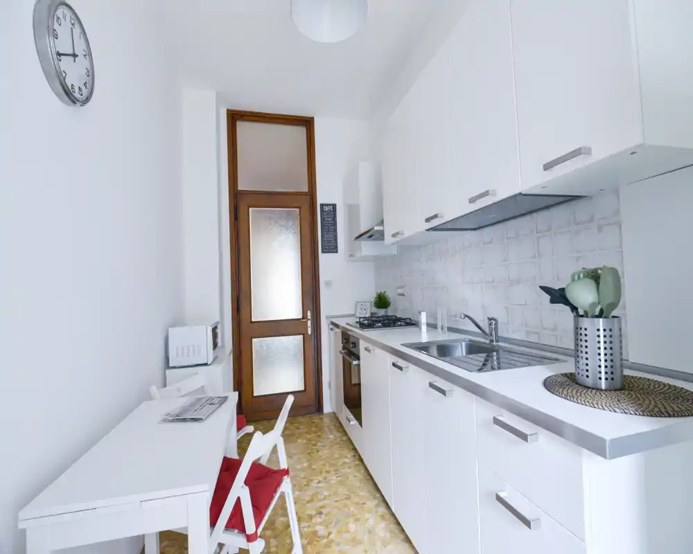 Cocina moderna y luminosa en disposición lineal con armarios blancos, fregadero de acero, horno encastrado y pequeña mesa — ideal para un piso compacto.