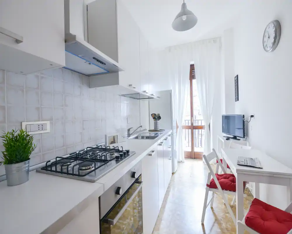 Cocina moderna y luminosa tipo pasillo con placa de gas, horno empotrado, fregadero y pequeña zona de comedor junto a la puerta del balcón. Superficies blancas y decoración sencilla, ideal para alquileres temporales.