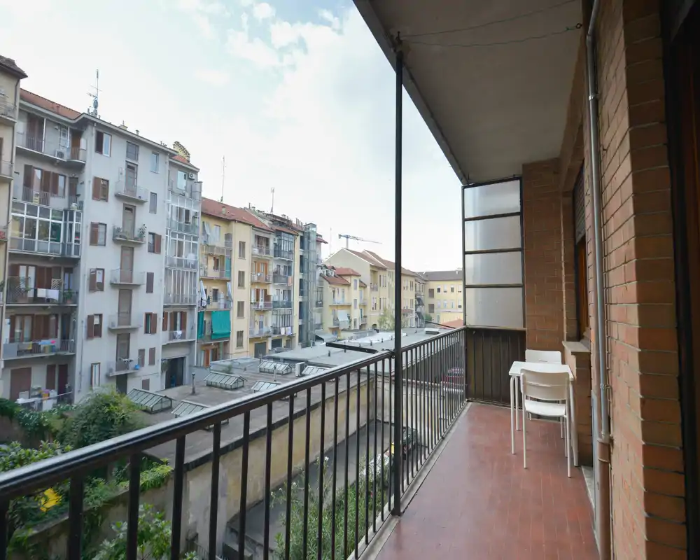 Balcón soleado con suelo de baldosas, barandilla metálica y una pequeña mesa con sillas con vistas a los edificios vecinos — ideal para el café de la mañana.