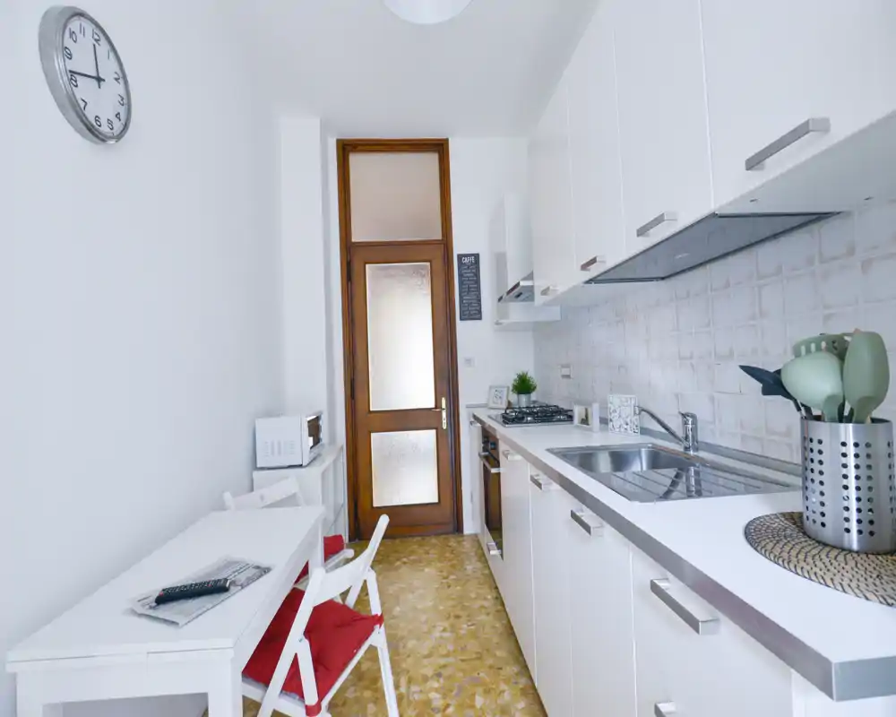Cocina en línea luminosa y moderna con armarios blancos, fregadero de acero, placa de gas y pequeña mesa con cojines rojos — limpia y bien presentada para un anuncio.