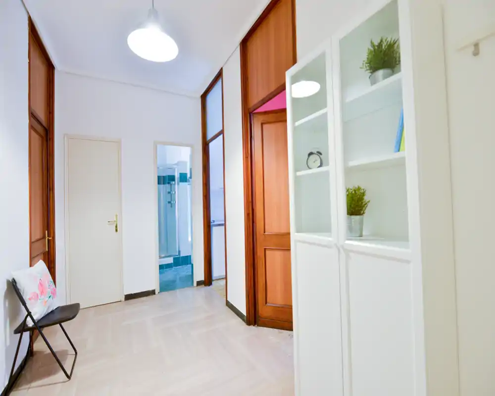 Recibidor luminoso y acogedor con paredes blancas, puertas de madera y un armario estrecho; suelo limpio y una silla plegable que ofrecen una primera impresión ordenada y funcional.