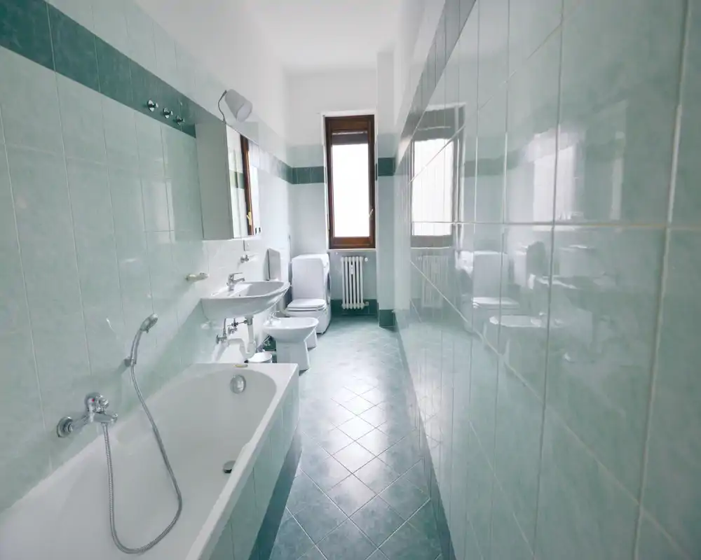 Baño luminoso con azulejos, bañera, lavabo, bidé e inodoro alineados en una pared y una ventana al fondo que aporta luz natural.