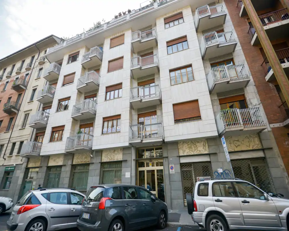 Fachada a nivel de calle de un edificio residencial de varias plantas con balcones y coches aparcados delante, adecuada para un anuncio que muestre la fachada y la entrada.