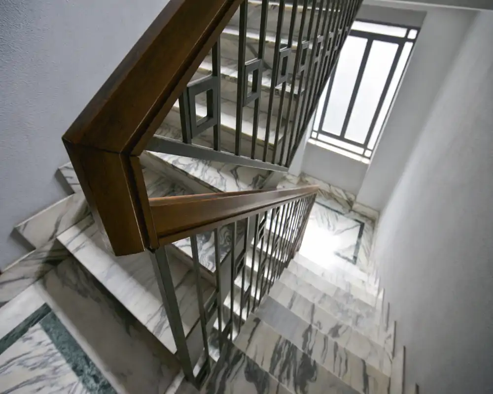 Una escalera de mármol bien iluminada con barandillas metálicas y pasamanos de madera, vista desde arriba. Escalera moderna y limpia, adecuada para la entrada de un edificio o un hueco interior.