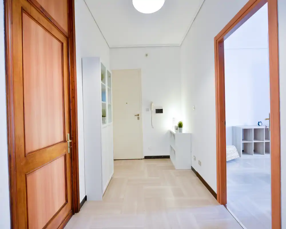 Entrada luminosa y ordenada con suelo de madera clara, un mueble blanco para almacenaje y una puerta hacia la habitación contigua.
