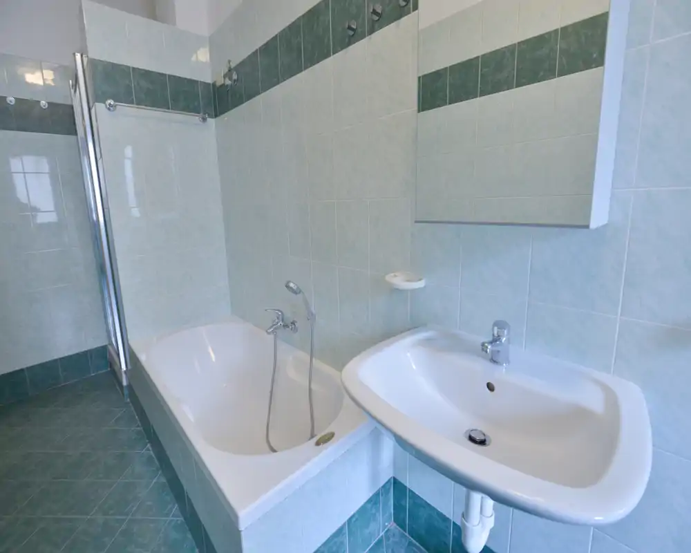 Baño limpio con paredes alicatadas, una bañera con ducha de mano y un lavabo de pared con armario espejo arriba; azulejos verdes neutros y buena luz natural.