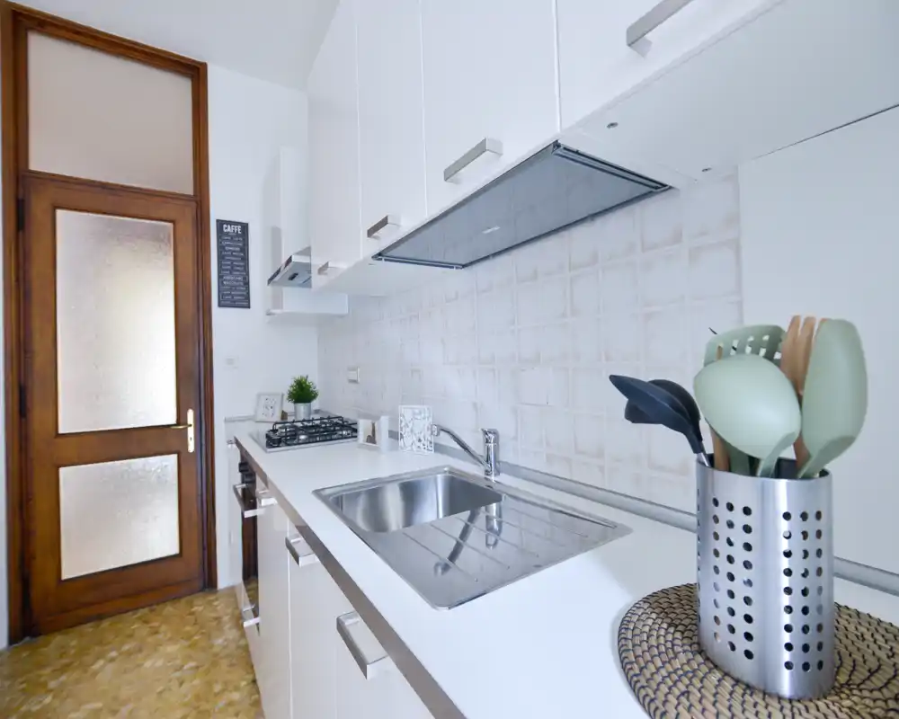 Cocina tipo pasillo, moderna y luminosa con muebles blancos, fregadero de acero y placa de gas. Detalles sencillos como utensilios y una planta pequeña transmiten un espacio funcional y cuidado.
