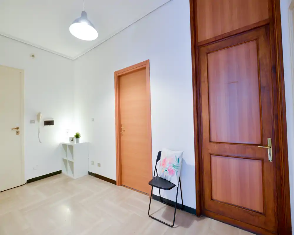 Entrada/pasillo luminoso y minimalista con dos puertas, una pequeña estantería y una silla plegable — paleta neutra y luz suave superior, ideal como zona de bienvenida.